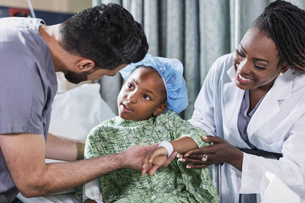 Ein sechsjähriges afroamerikanisches Mädchen sitzt auf einer Krankenhausliege und wird von einem Arzt und einer Krankenschwester versorgt.  Sie trägt einen Krankenhauskittel und eine chirurgische Kappe. Auf der einen Seite steht ein Krankenpfleger im OP-Kittel, ein junger Mann aus dem Nahen Osten in seinen Zwanzigern, und auf der anderen Seite steht eine Ärztin, eine Afroamerikanerin in ihren Vierzigern.