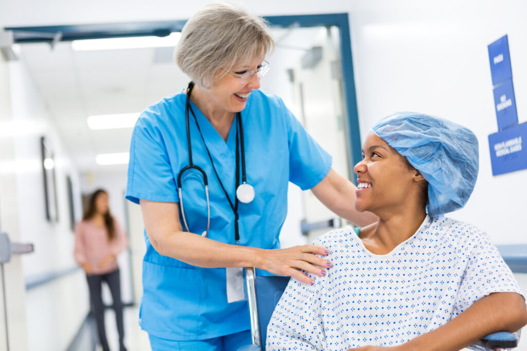 Eine ältere Krankenschwester mit grauem Haar schiebt eine Patientin im Rollstuhl. Sie betreut eine Patientin, die sich nach einem ambulanten chirurgischen Eingriff erholt. Bei der Patientin handelt es sich um eine junge Afroamerikanerin, die ein Krankenhaushemd und eine OP-Haube trägt. Die Frauen lächeln, während sie miteinander sprechen.